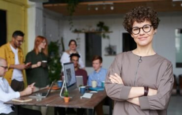 Smiling businesswoman with curly hair stands confidently in a modern office space with colleagues.